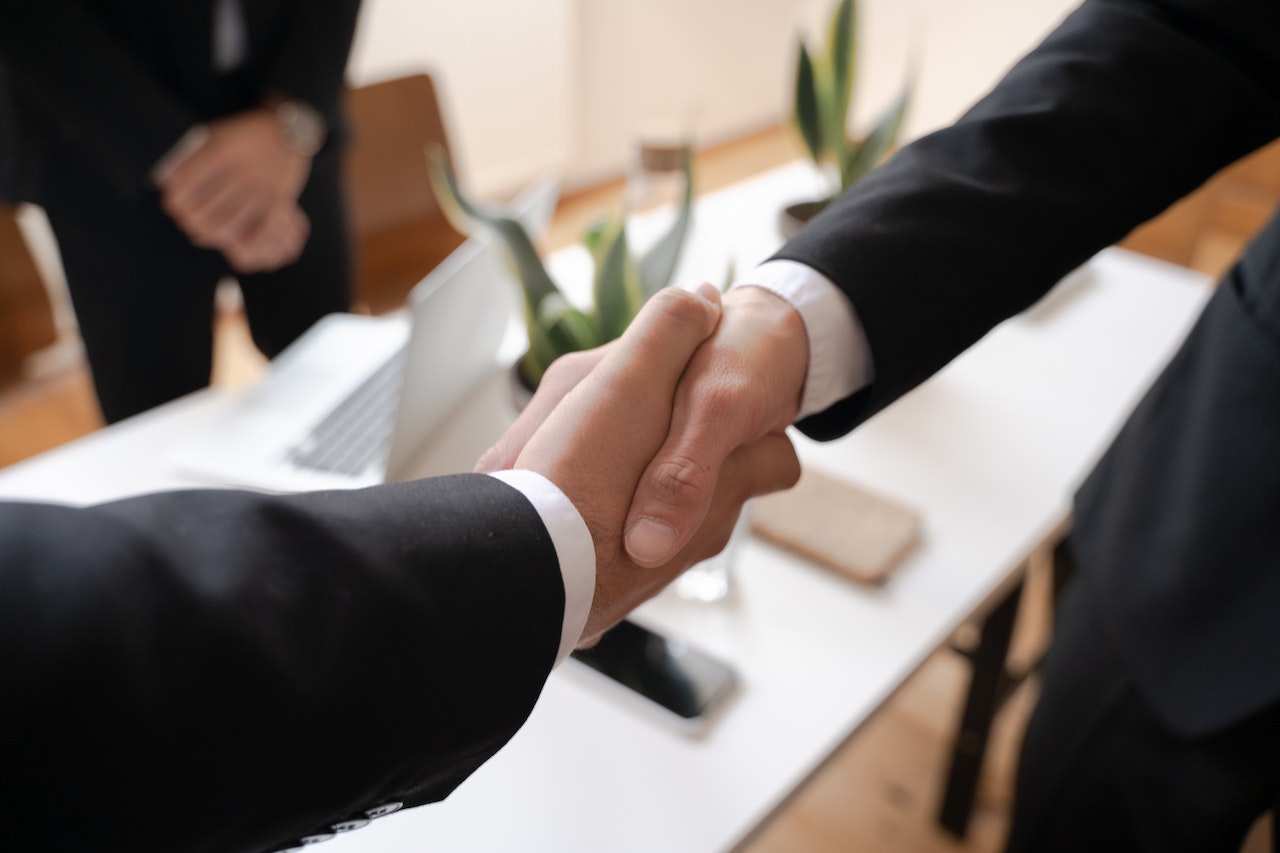 Two people shake hands in close-up view, their hands clasped together in a firm grip. Both individuals are dressed in professional attire and appear to be engaged in a friendly and positive interaction